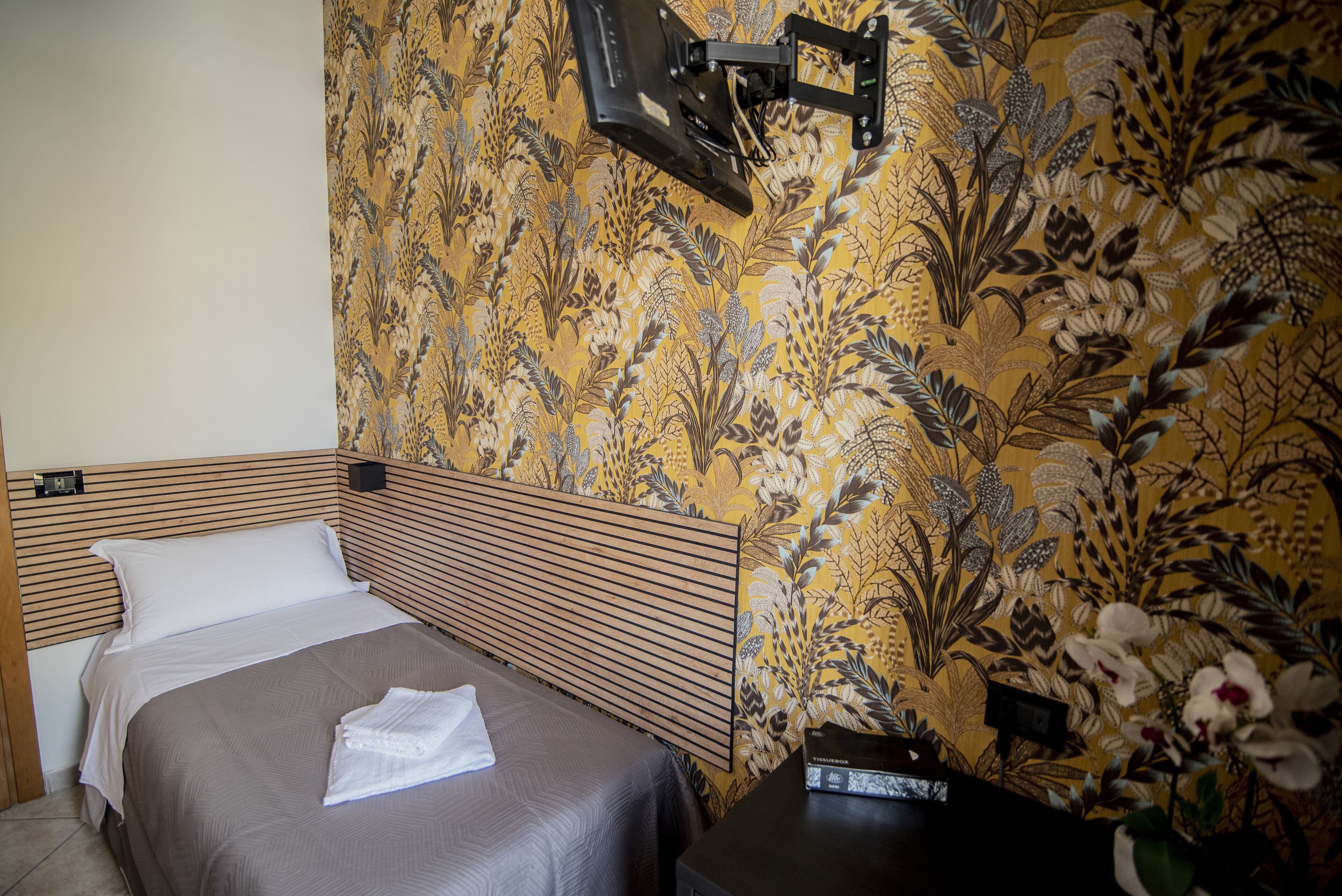 A hotel room with a bed, a nightstand, a wall-mounted TV, and a floral wallpaper.