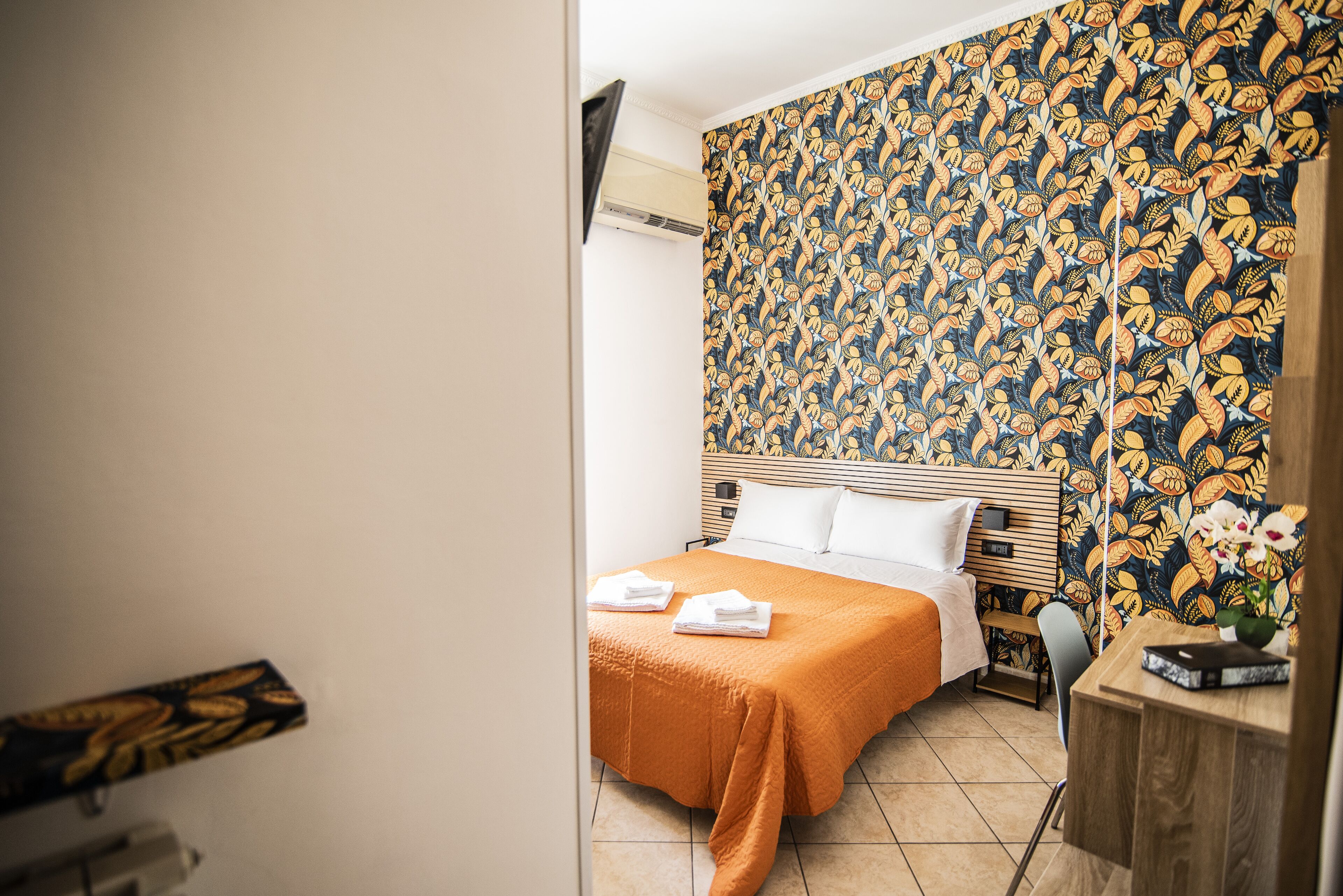 A hotel room with a bed, a desk, and a patterned wallpaper.