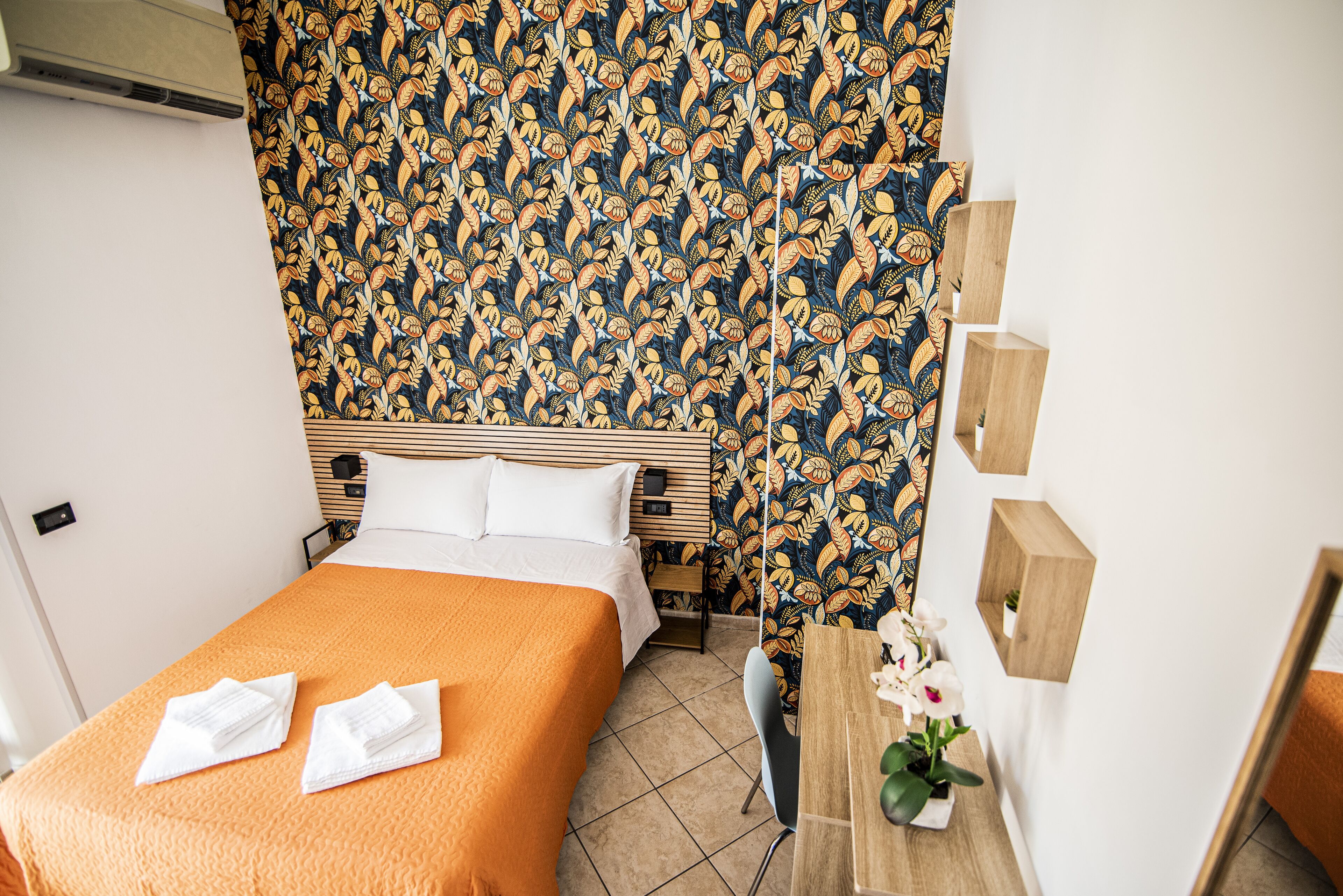 A hotel room with a bed, a wooden desk, a chair, and a wall with a floral pattern.
