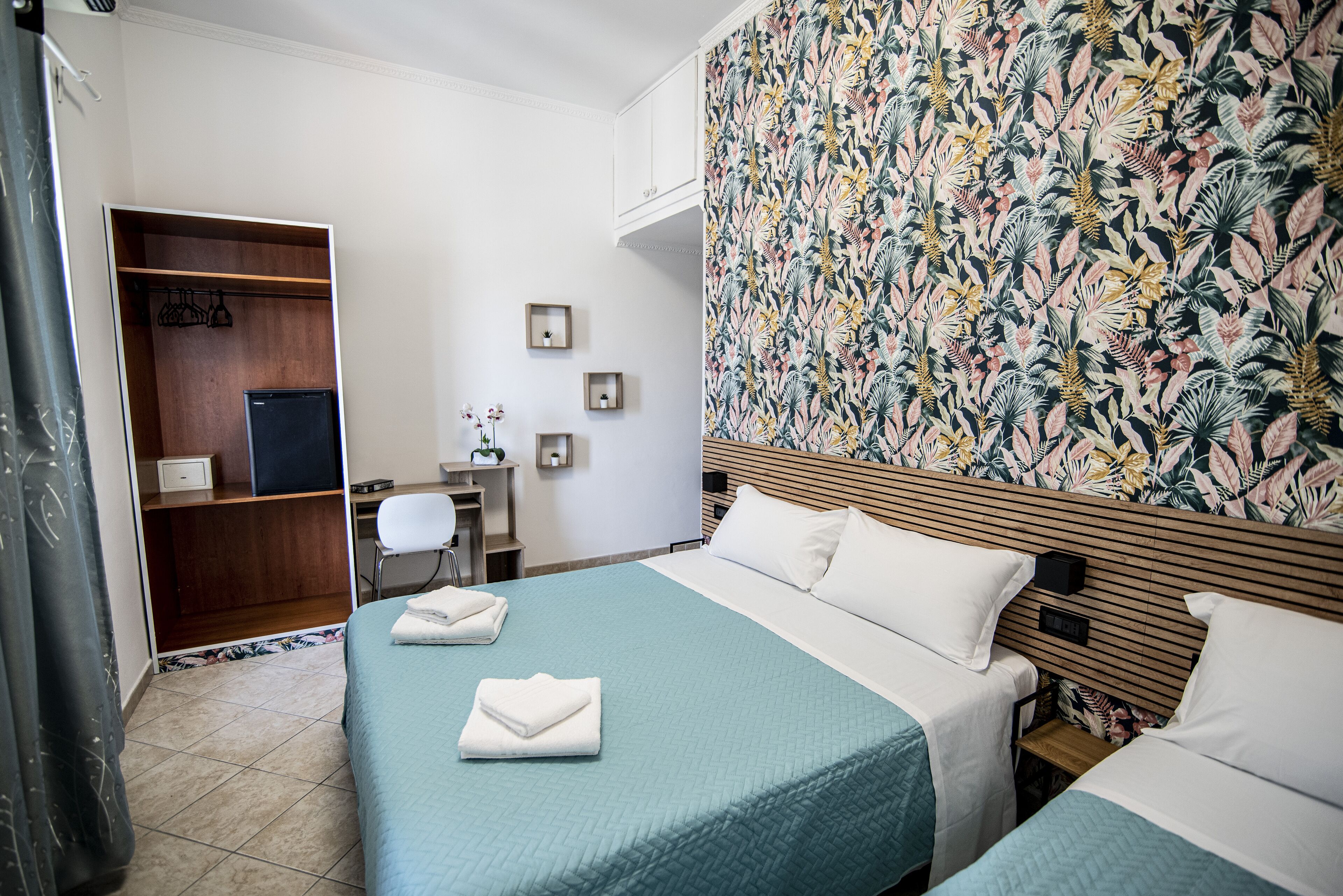 A hotel room with two beds, a wooden wardrobe, a desk with a chair, and a floral-patterned wall.