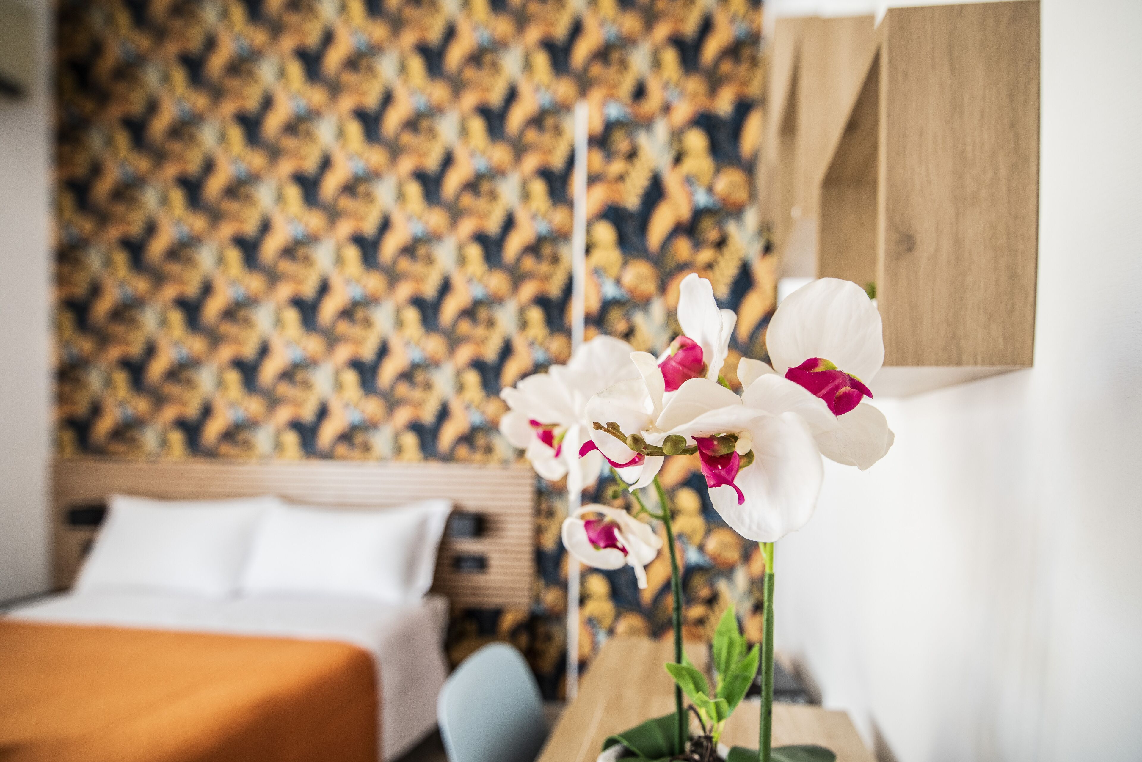 Une chambre d’hôtel avec un papier peint à motifs, un lit avec une literie blanche et une plante orchidée en pot.