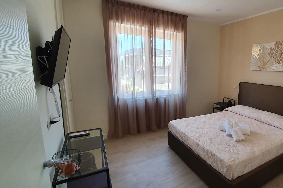 Une chambre à coucher avec un lit, une table de chevet, un téléviseur fixé au mur, une fenêtre avec des rideaux et un tableau.