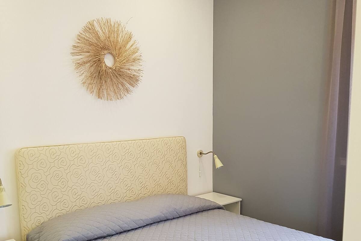 Un lit bien fait, avec une couverture matelassée grise et des oreillers, une table de chevet avec une lampe, et un objet décoratif fixé au mur.