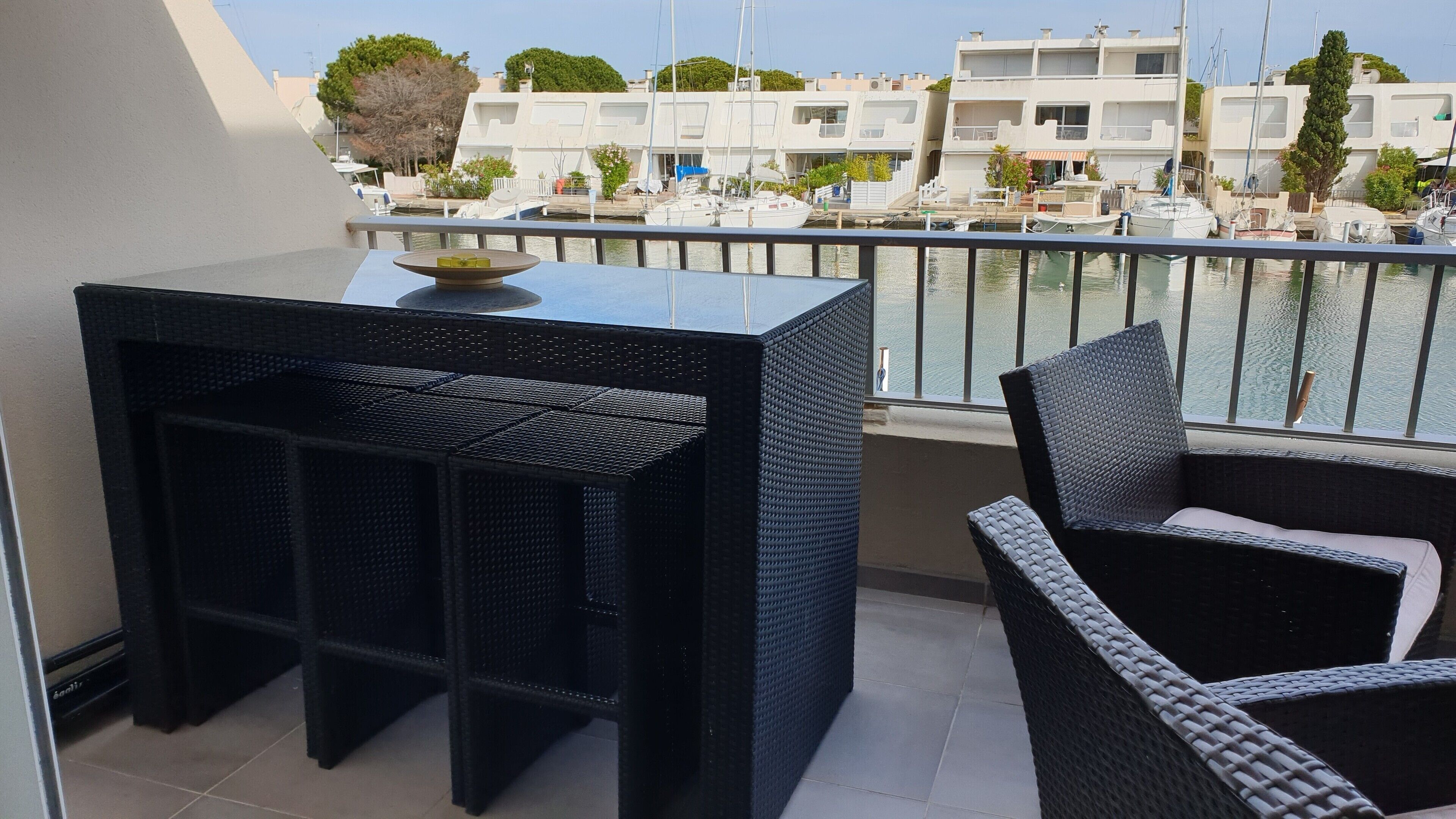A balcony with a black wicker table and chair, overlooking a marina with boats and buildings.