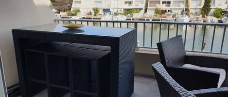 A balcony with a black wicker table and chair, overlooking a marina with boats and buildings.