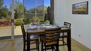 Zona pranzo con un tavolo grande apparecchiato per quattro persone, una porta scorrevole in vetro che dà su un patio con vista sulle montagne e un quadro di un paesaggio appeso al muro.