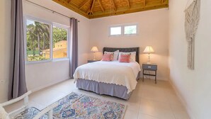 A bedroom with a large bed, two bedside tables with lamps, a window with curtains, and a macrame wall hanging. - Hospitality Expert CP (Montego Bay)