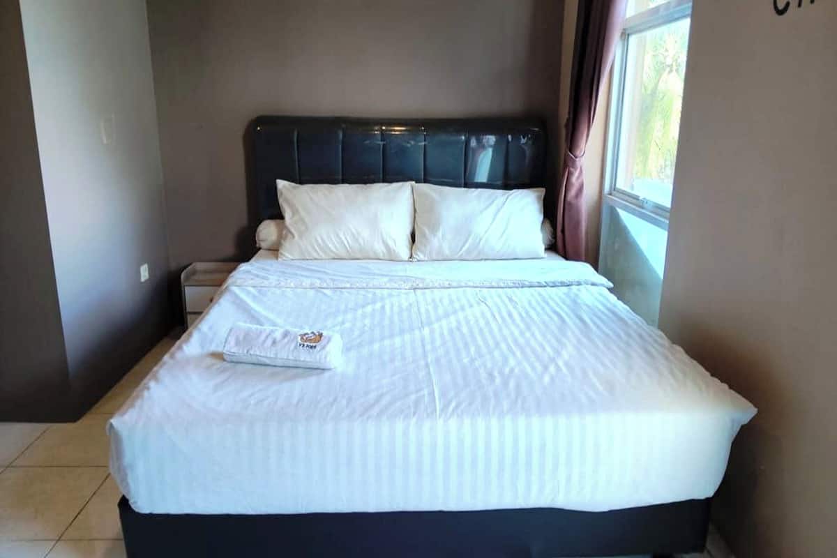 Una cama bien hecha con sábanas blancas y un cabecero negro, una mesita de noche con una lámpara, y una ventana con cortinas.