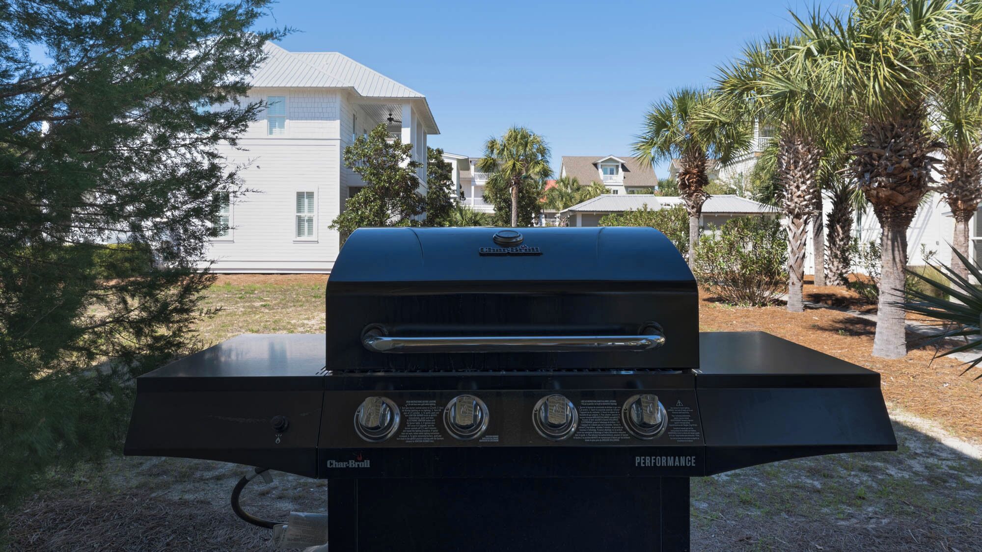 A Char-Broil grill with four burners, set outdoors with palm trees and houses in the background.