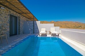A pool with two lounge chairs, a stone wall, and a view of mountains.