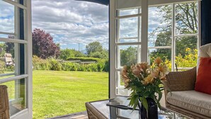 A room with a glass table, wicker chairs, a sofa, and a view of a well-maintained garden through large windows. - 2 bedroom accommodation in Richards Castle, near Ludlow (Richards Castle, near Ludlow)