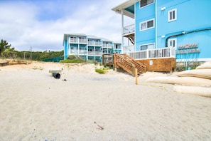 Un hôtel situé en bord de mer, doté d’un escalier en bois menant à un balcon, d’une plage de sable et d’un ciel dégagé.