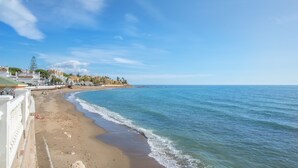 Una playa costera con un paseo marítimo, cielos despejados y edificios al fondo.