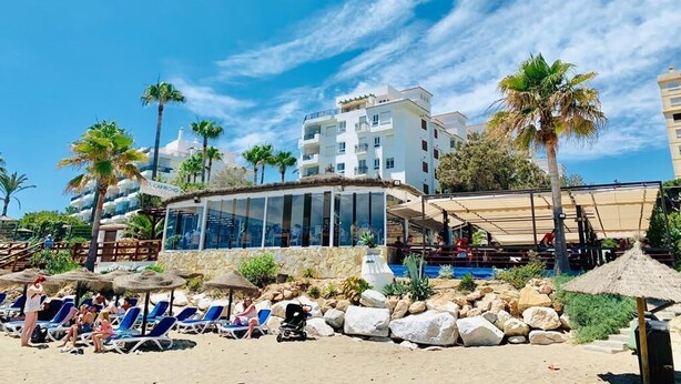 Un hotel frente a la playa con piscina, tumbonas y palmeras.