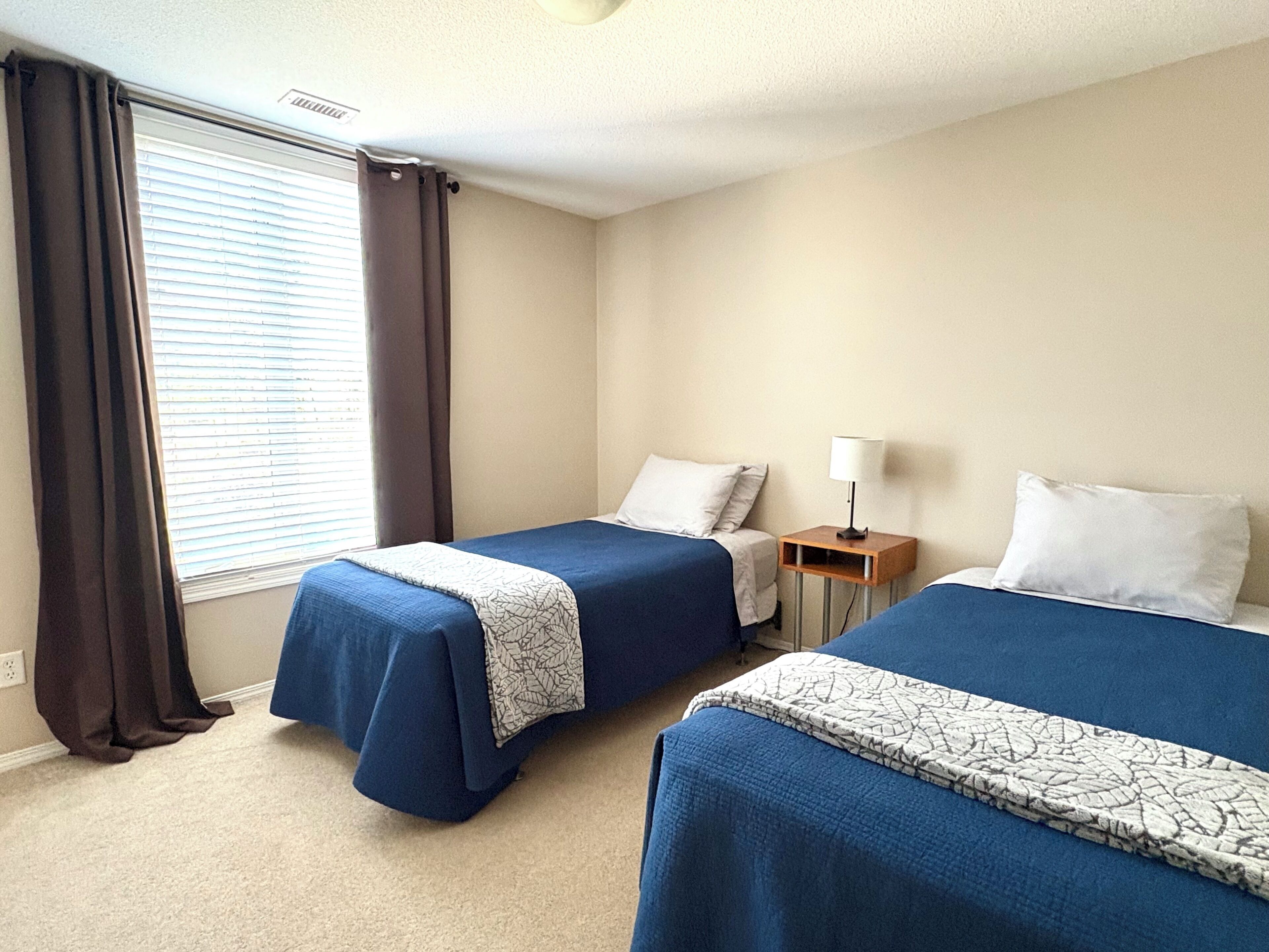 A hotel room with two beds, a bedside table with a lamp, a window with curtains, and a carpeted floor.