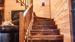 Una escalera de madera en un interior tipo cabaña, con un sofá de cuero y un mapache asomándose por una ventana.