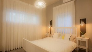 Un dormitorio con cama, cortinas blancas, mesita de noche, lámpara y aplique de pared.