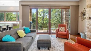 A modern living room with a grey sofa, an ottoman, and a red armchair. There is a stone wall and a large window with a view of trees. - Casita Grove-2 Lakeside Homes w/Casita & Boat Lift (Burnet)