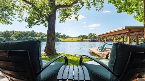 A covered outdoor seating area with two chairs and a table, overlooking a lake with a dock and a covered structure in the background. - Casita Grove-2 Lakeside Homes w/Casita & Boat Lift (Burnet)