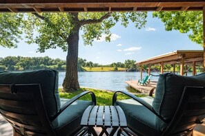 A covered outdoor seating area with two chairs and a table, overlooking a lake with a dock and a covered structure in the background. - Casita Grove-2 Lakeside Homes w/Casita & Boat Lift (Burnet)