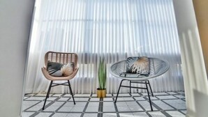 Una habitación con piso de baldosas, dos sillas de mimbre, una planta en maceta y una ventana con persianas verticales.
