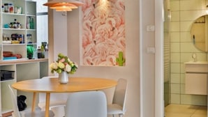 Un comedor con una mesa redonda, sillas, un florero, una pared con un mural de flamencos y una zona de cocina con estanterías.