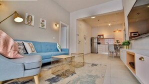Una sala de estar moderna con un sofá azul, una mesa de centro y una alfombra. Se ve una zona de cocina al fondo.