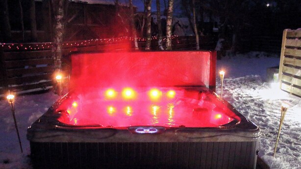 A hot tub with lights, surrounded by snow, and a wooden fence in the background.