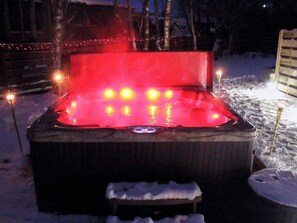 Uma banheira de hidromassagem com luzes, rodeada por neve, e um cercado de madeira ao fundo.