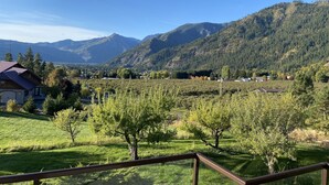 A mountainous landscape with a clear sky, a wooden deck, and a view of a valley with scattered houses and trees. - Ski Hill home w/ stunning views & hot tub! (Leavenworth)