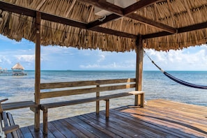 A wooden deck with a thatched roof, a hammock, and a bench overlooking a calm sea.
