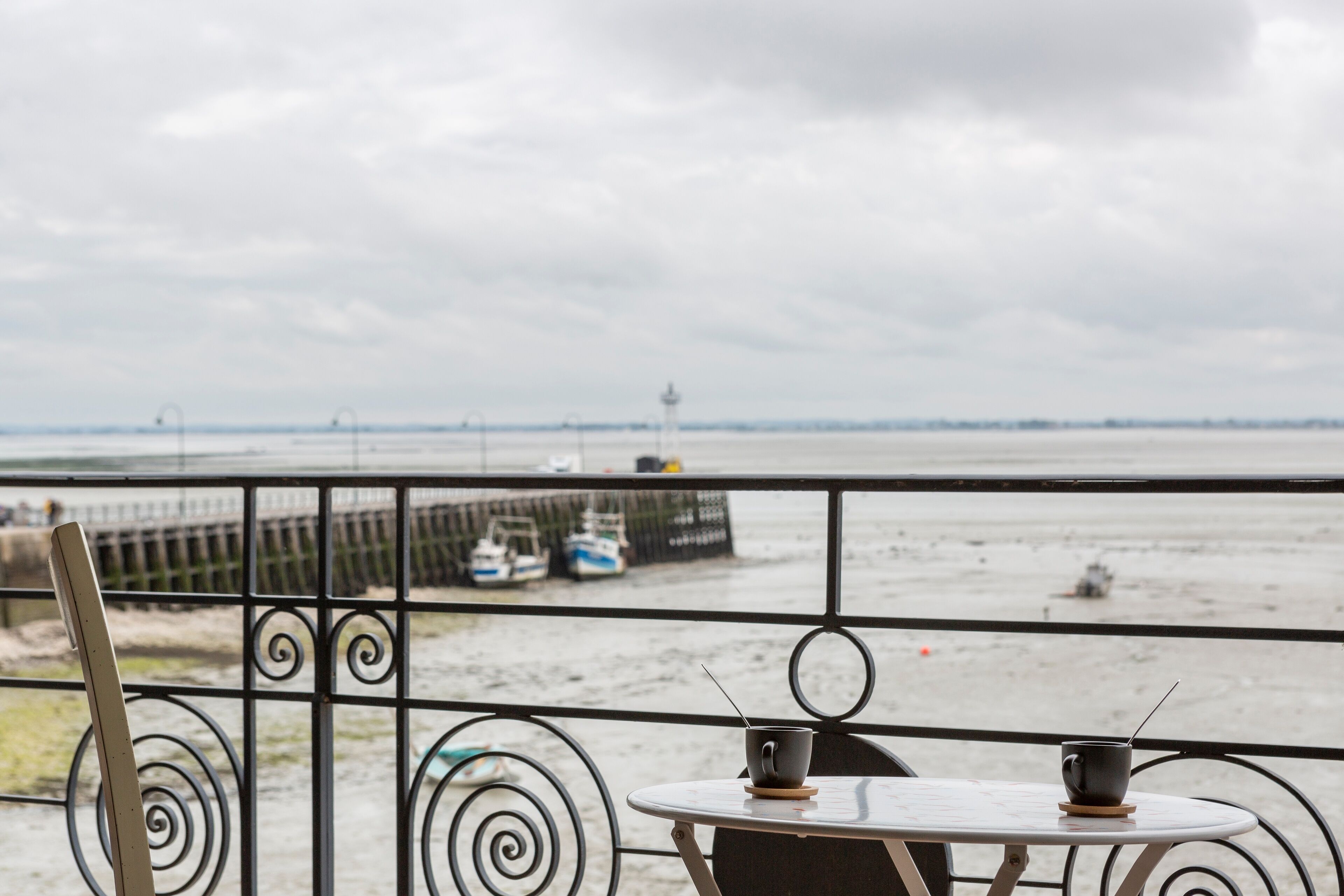 A balcony with a table and chairs, overlooking a beach with boats and a pier.