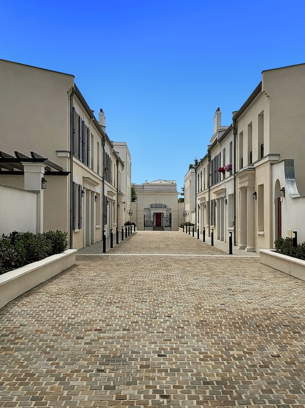 A cobblestone pathway leading to a building with a red door, flanked by white buildings with black shutters.