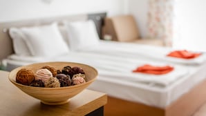 A bed with white bedding and a wooden nightstand with a bowl of assorted chocolates. - Step (Sunny Beach)