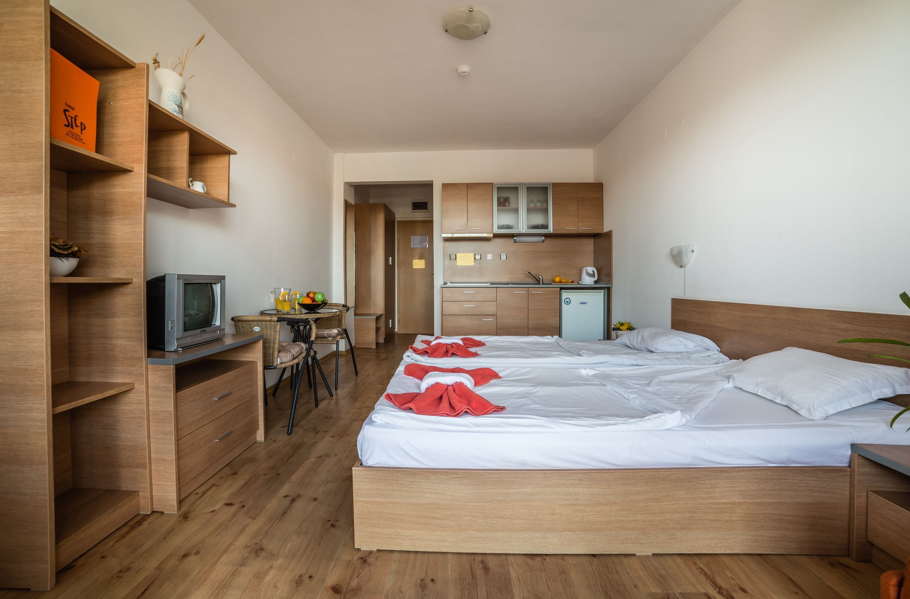 Une chambre d’hôtel avec un lit en bois, un bureau avec une chaise, un coin cuisine et un téléviseur posé sur une étagère en bois.