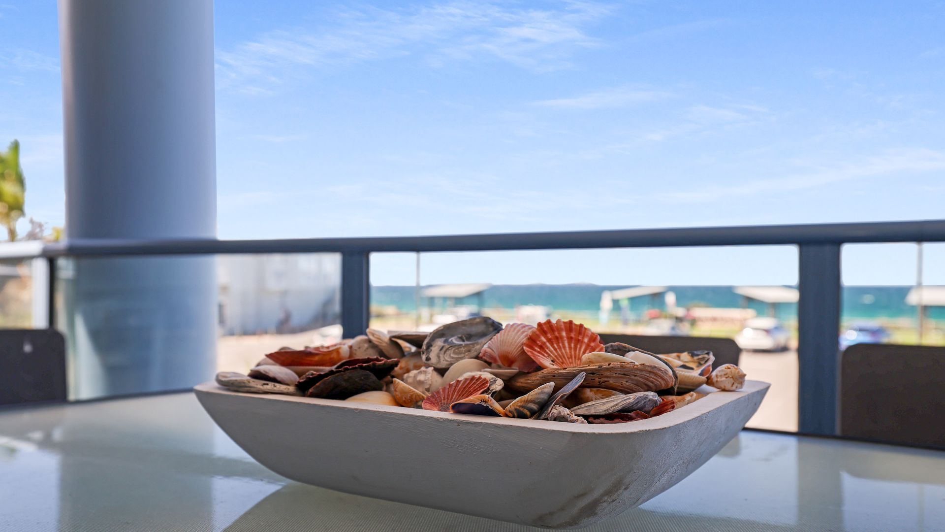 A bowl of seashells on a table with a view of the sea and buildings in the background.