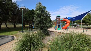 A playground with a slide and climbing structures, surrounded by trees and a clear sky. - Enjoy the soothing sea breeze (Hawks Nest)