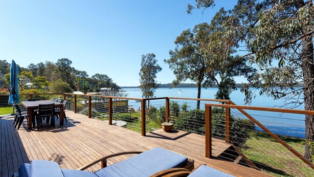 A wooden deck with a table and chairs, overlooking a lake with a boat in the distance.