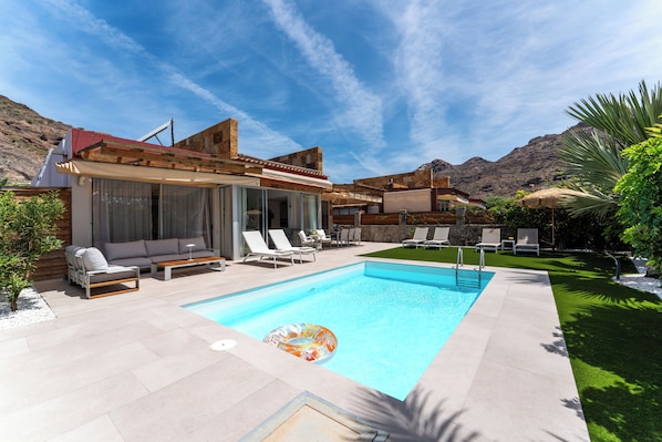 A swimming pool with lounge chairs and a sun umbrella, surrounded by a landscaped garden and a mountain backdrop.