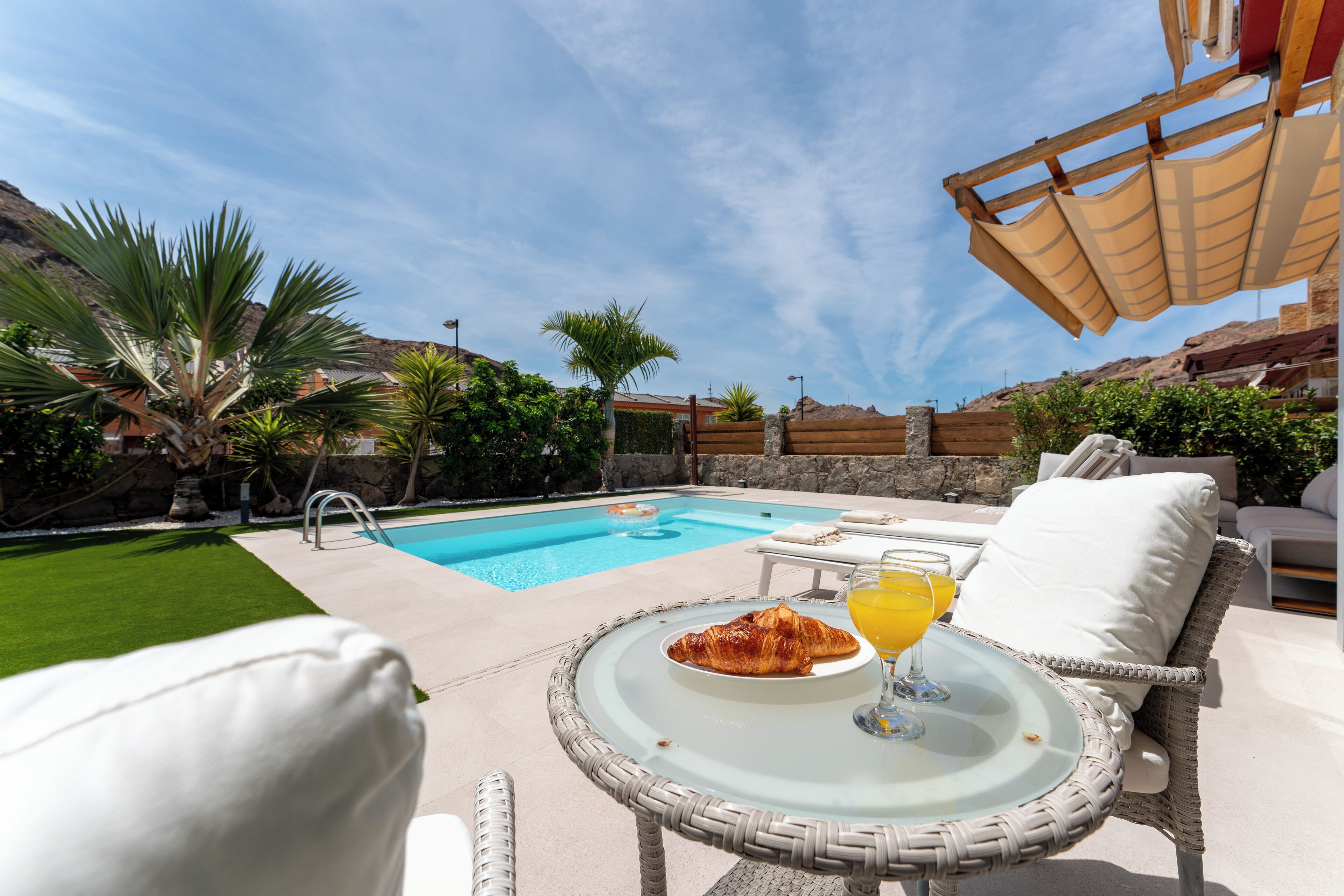 A poolside setting with a dining table, a glass of orange juice, and a croissant.