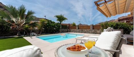 A poolside setting with a dining table, a glass of orange juice, and a croissant.