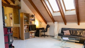Een loft-achtige kamer met een schuine plafond, een glazen salontafel, een leren bank, een houten boekenplank, en een bureau met stoel.