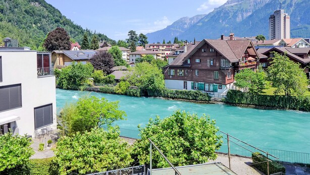 Een hotel aan de rivier met traditionele architectuur, uitzicht op de rivier en bergen op de achtergrond.