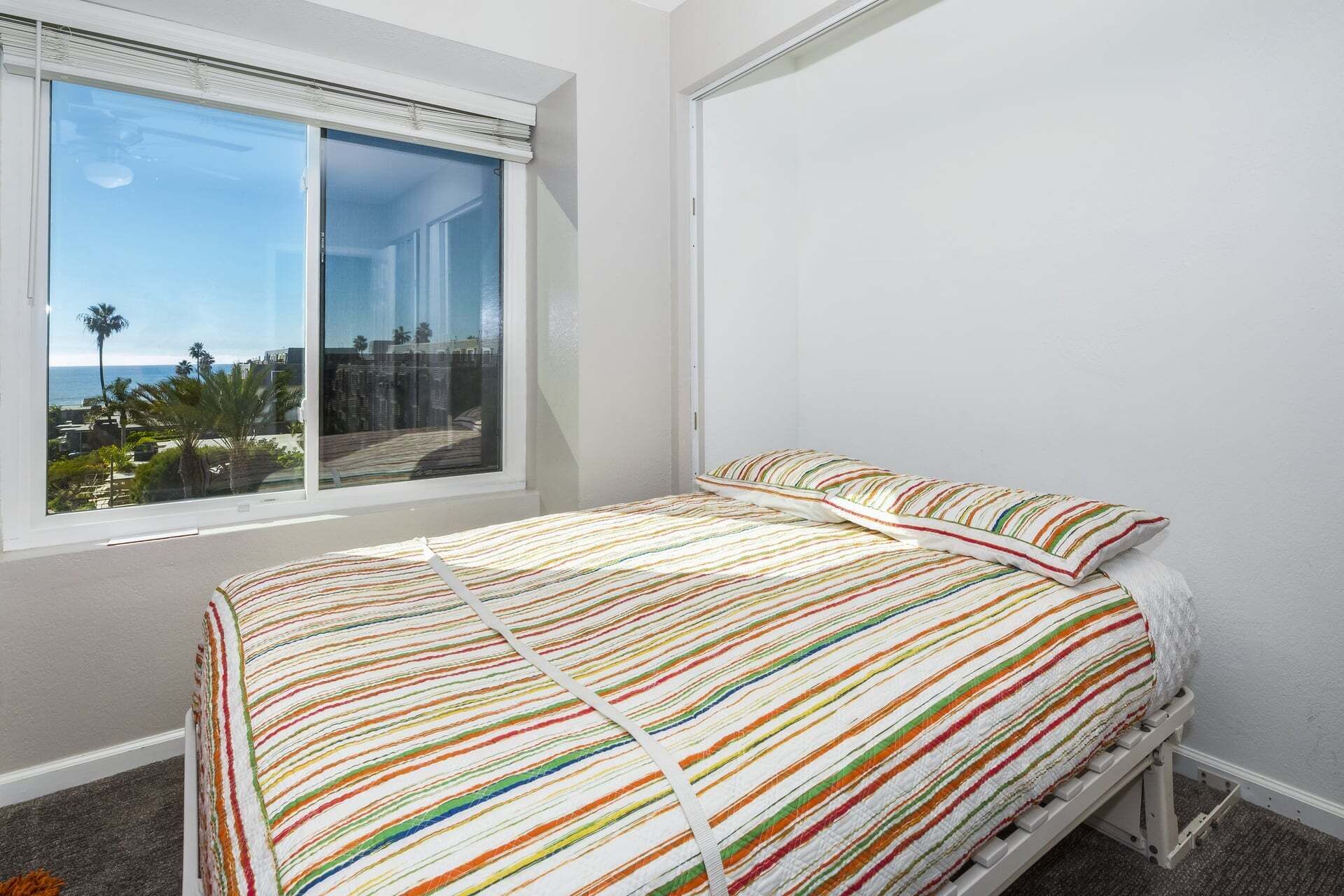 A bedroom with a bed, a window with a view of palm trees and buildings, and a white wall.