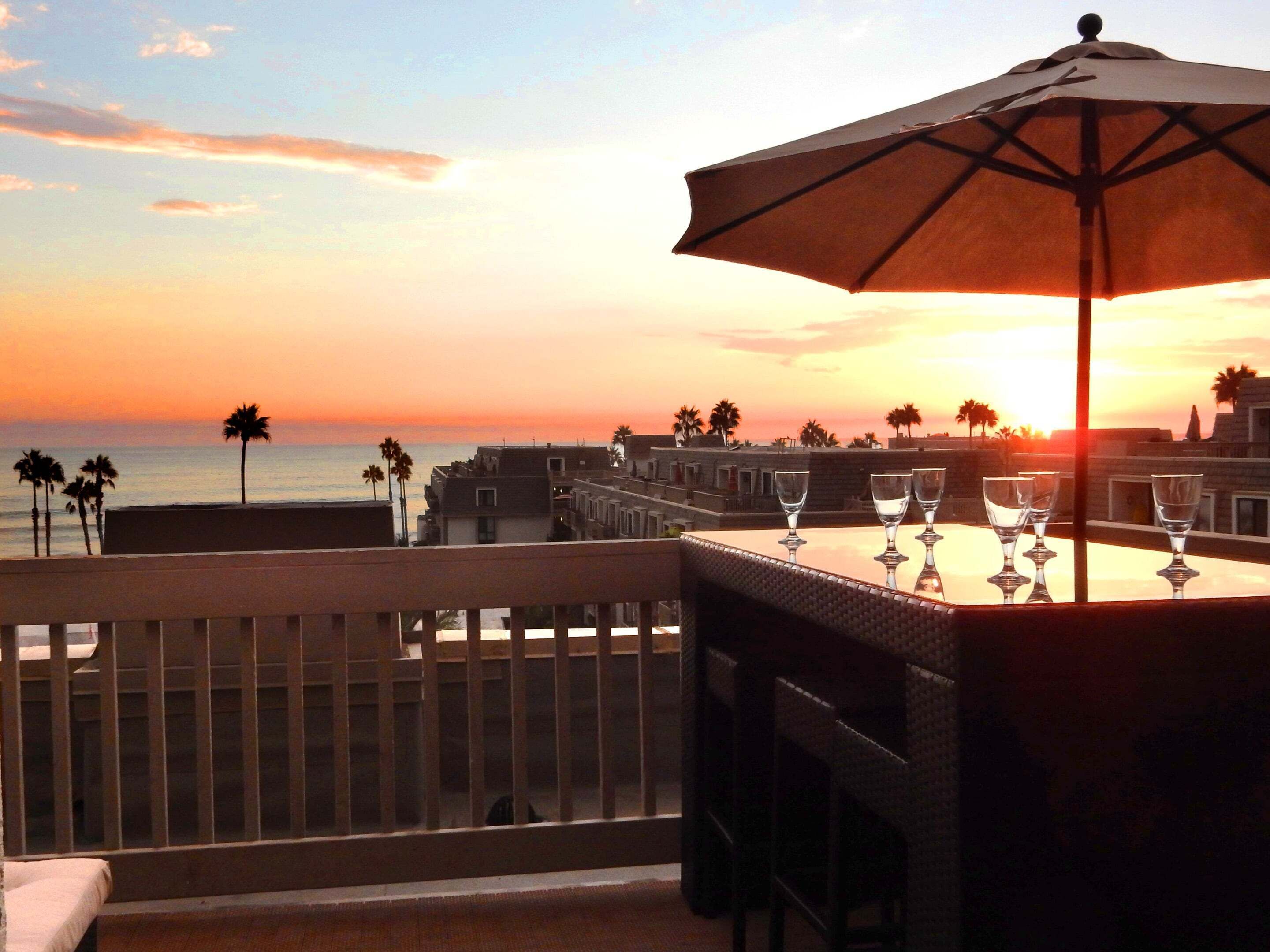 Un balcon avec une table et des chaises, offrant une vue sur le coucher de soleil, avec des palmiers et des bâtiments en arrière-plan.