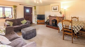 A living room with a sofa, ottoman, television, fireplace, and a dining table with chairs. - Trewin Court - UK11878 (Holsworthy, near Launceston)