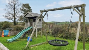 A wooden play structure with a slide and swings, surrounded by grass and trees. - Trewin Court - UK11878 (Holsworthy, near Launceston)