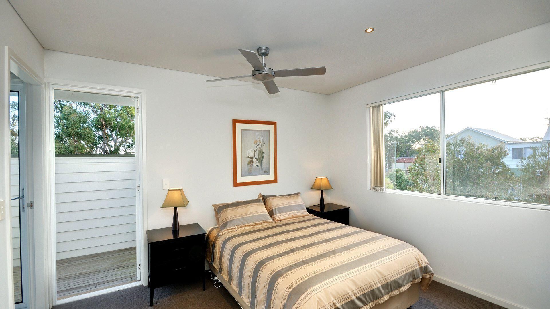 A bedroom with a bed, two bedside tables with lamps, a ceiling fan, a window with a view, and a door leading to an outdoor area.