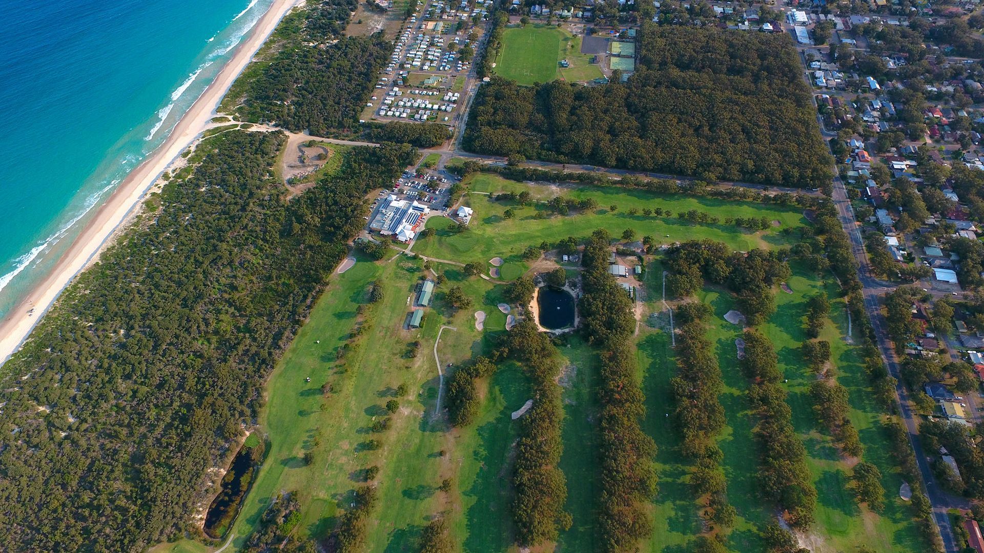 An aerial view of a coastal area with a beach, dense forest, and residential buildings.