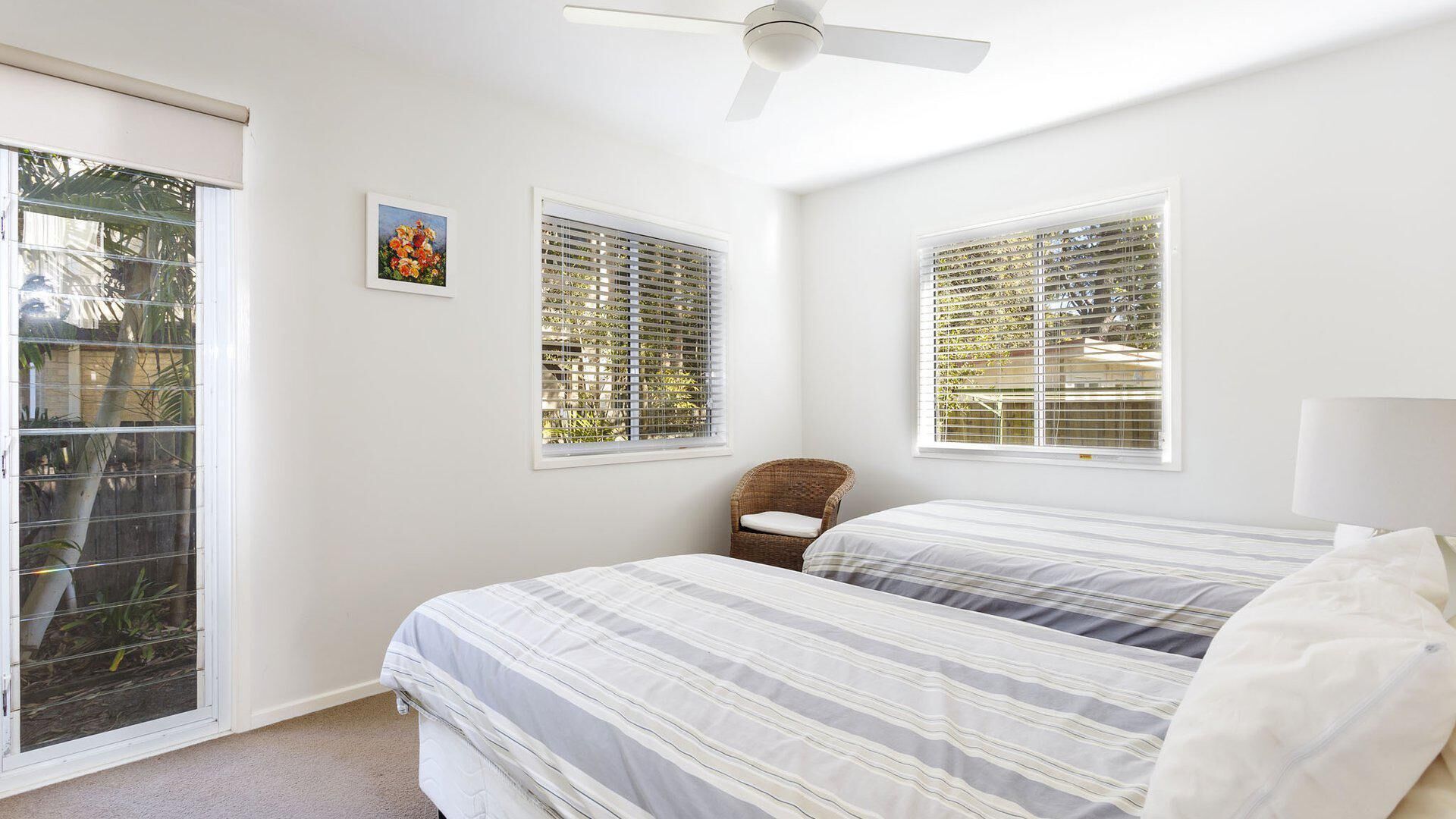 Ein Schlafzimmer mit zwei Betten, einem Deckenventilator und großen Fenstern mit Jalousien.